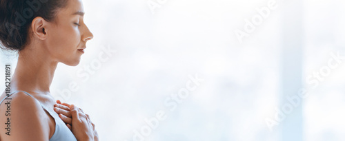 Fotografie A woman stands in a well-lit room with her eyes closed and hands on her chest