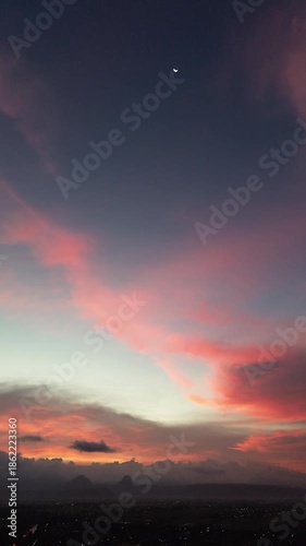 Cinematic aerial footage of colorful sunrise sky with red and orange clouds and a crescent moon above a dark landscape. Peaceful, dramatic atmosphere ideal for background and cinematic visuals.