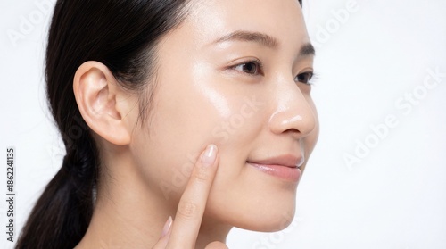 Profile of young Asian woman showcasing radiant, glass skin, gently touching her moisturized cheek, highlighting the glossy effect of premium beauty and cosmetic treatment.
