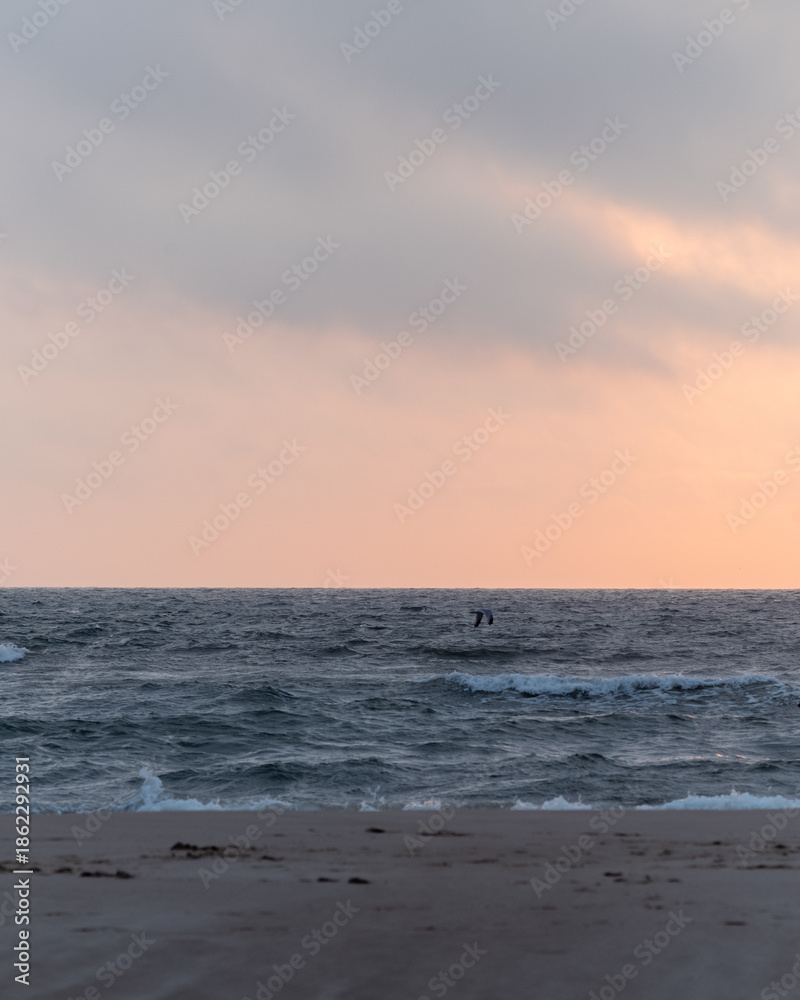 Obraz premium Waves gently crash on a quiet beach at sunset with a soft pink sky and distant figure in the water