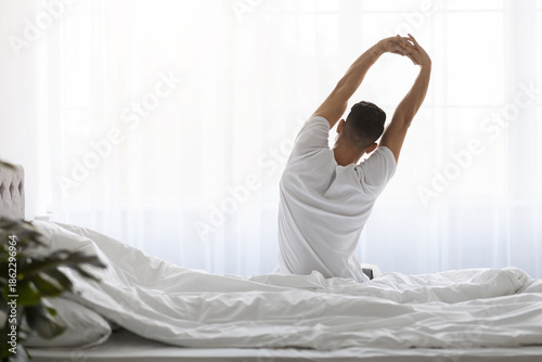 Slika na platnu Young man sits on his bed with his back facing the viewer, stretching his arms high after waking up