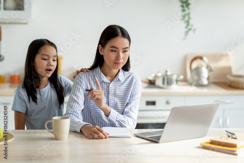 Bored asian girl demanding attention from her busy mother, woman working at home and ignoring her lonely child. Freelancer lady using laptop while sitting in kitchen, copy space