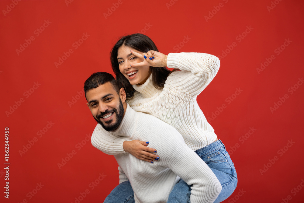 Obraz premium A joyful Arab couple enjoys a playful moment together. The man gives his girlfriend a piggyback ride as she poses with a peace sign. They are clearly having fun against a vibrant red background.