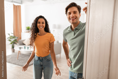 Fotografie Cheerful young couple stands in the doorway of their modern flat, inviting guests inside with warm smiles