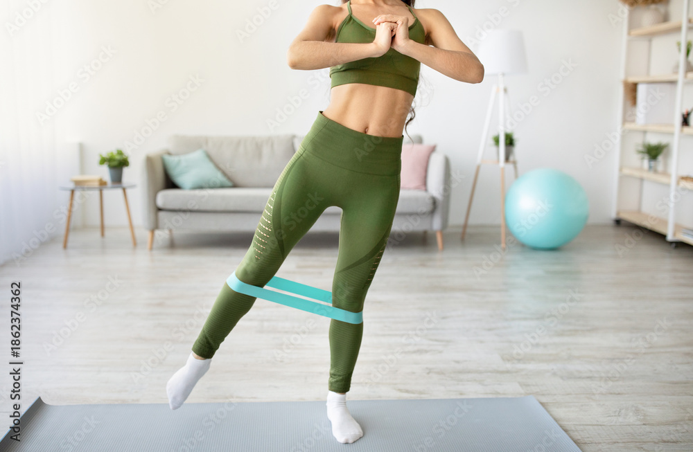 Obraz premium Unrecognizable young lady exercising with elastic band, working out her strength in living room. Motivated millennial woman staying fit and healthy at home during covid-19 lockdown, closeup