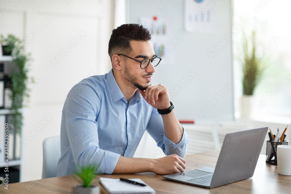 Fototapeta premium Positive Arab businessman using laptop pc at workplace, speaking to colleagues on webcam. Successful millennial office worker reading important document online, checking email