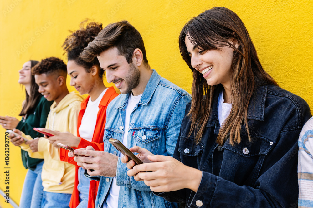 Obraz premium Happy group of young multiracial friends using smart phones together outside over yellow background. Social media and technology lifestyle concept