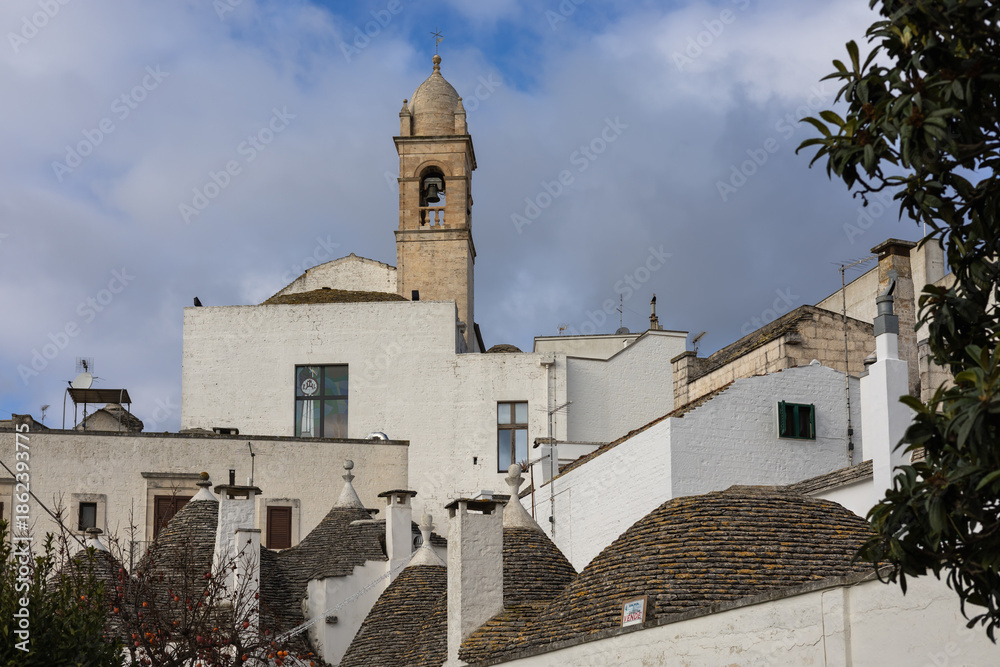 Fototapeta premium traditional trulli houses with conical stone roofs and white walls under a bell tower in alberobello, puglia, italy. authentic mediterranean village.