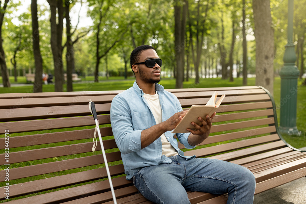 Obraz premium Young black visually impaired man sitting on bench in city park, reading Braille book outdoors. Millennial blind guy in dark glasses studying or enjoying entertainment literature, outside