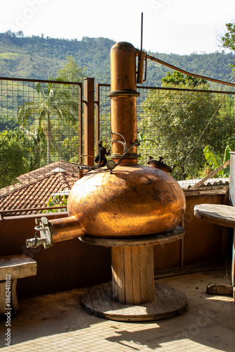 Tableau sur toile Traditional copper alembic still for distillation, set against a backdrop of lush green hills and tropical foliage, suggesting artisanal production