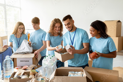 Slika na platnu Group of young volunteers from diverse backgrounds pack canned food and other products into cardboard boxes for a charitable foundation