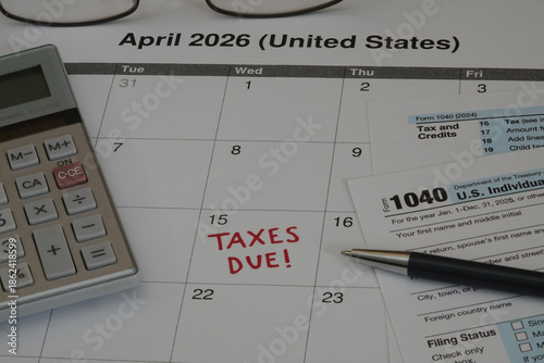 A 2026 calendar noting the April 15 USA Internal Revenue Service IRS income filing deadline for year 2025 taxes is shown up close, with a calculator, ink pen, and glasses in the frame.