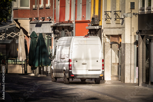 A white delivery van driving down a paved urban street lined with shops in the city of Heerlen, The Netherlands, a symbol of business and logistics, last mile delivery.