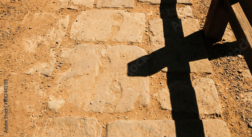 Shadow of wooden cross cast on ancient dusty stone path with footprints representing concept of following Jesus