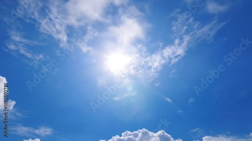 A serene and expansive blue sky dominates the frame, showcasing a beautiful interplay of white clouds, ranging from wispy cirrus to soft, fluffy cumulus formations. The brilliant sun shines brightly, 