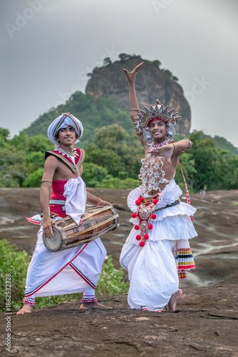 SIGIRIYA, SRI LANKA - DECEMBER 04, 2025 : A Gatabera Player (Getaberakaruwo) and a Ves Dancer or Up Country Dancer perform in front of Sigiriya Rock in Sri Lanka.