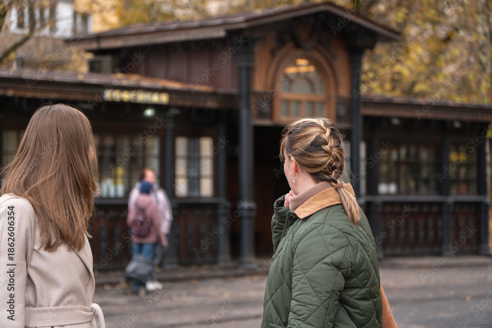 Fototapeta premium Two women stand near historic wooden tram stop in Moscow, surrounded by autumn trees, warm yellow leaves, classic urban architecture