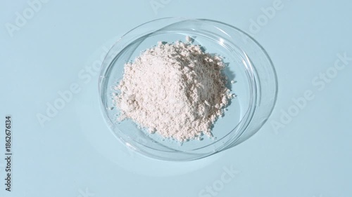 Vertical tracking shot of a stack of white fine powder inside a plastic petri dish on a pale blue background lit with orbiting light casting shifting shadows | Zinc stearate cosmetic ingredient