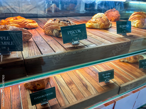 Fotografi Close-up of a wooden bakery display case featuring various labeled croissants and pastries including Piscok, Almond, Ham & Cheese, and Bi-Color varieties with elegant dark green price tags