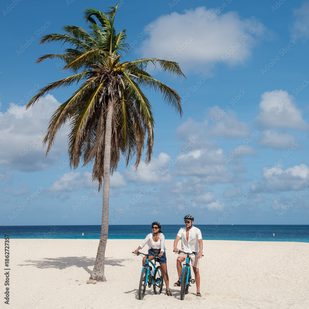 Naklejka premium Two cyclists enjoy a relaxing ride along the soft sands of Eagle Beach in Oranjestad, Aruba. The vibrant blue waters and lush palm trees create a perfect vacation atmosphere.