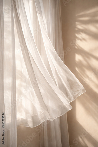 Φωτογραφία White sheer curtains gently blowing in sunlight with soft leaf shadows on a beige wall