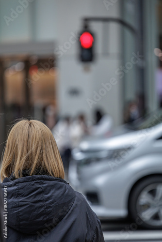 Candid urban street moment of blonde-haired women waiting for glowing red traffic light at busy crosswalk while car passes by in front of small group of people in bokeh background of gray winter day.
