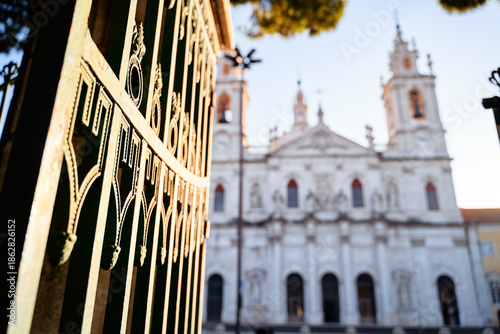 close-up view of an ornate green gate with intricate designs, leading to beautiful white church in the background. church features tall spires and detailed architecture, illuminated by soft sunlight.