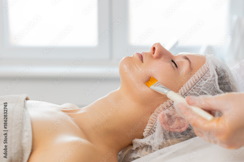 Fototapeta premium Caucasian young woman lies on couch while cosmetologist applies facial mask with brush on face in clinic. Relaxation skincare treatment gives smooth skin comfort wellness and calm mood during session.