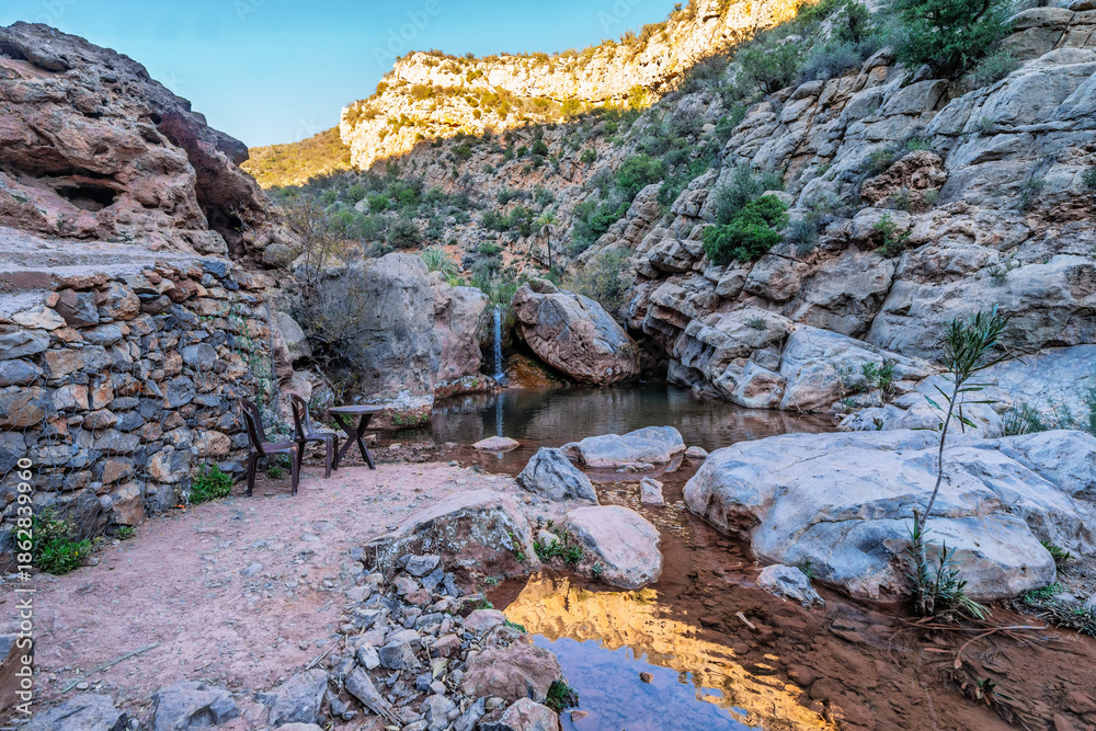 Fototapeta premium Serene Rocky Canyon Oasis With Pool, Trees, and Mountain Backdrop Under Blue Sky, Tizguan, Agadir, Morocco