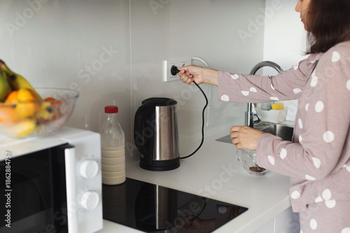 Foto Woman in polka dot pajamas plugging electric kettle into socket in home kitchen