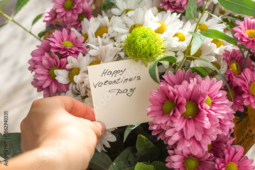 Romantic handwritten card with text happy valentine's day held in hand inside colorful bouquet of chrysanthemums and greenery, love message concept, valentines day greeting, celebration, affection