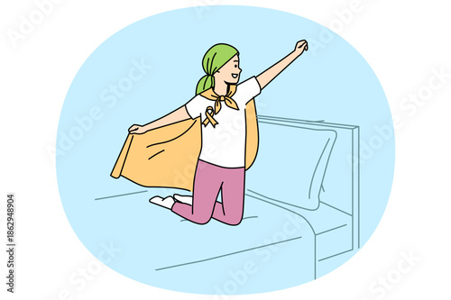 Child with oncology disease stands on bed in superhero pose, for concept day against cancer. Little boy with cancer feels improved mood after defeating deadly disease with chemotherapy