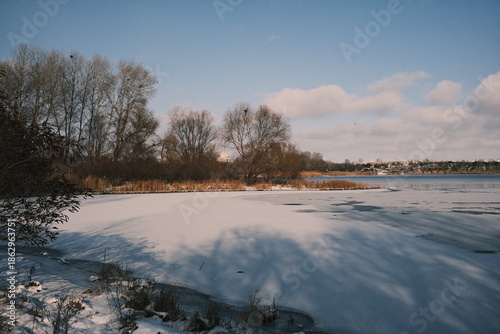Obraz na plátně Frozen lake landscape with snow-covered ice, reeds, and winter shoreline under a blue sky, calm seasonal scenery showing cold weather nature, tranquil water edges, and rural winter atmosphere