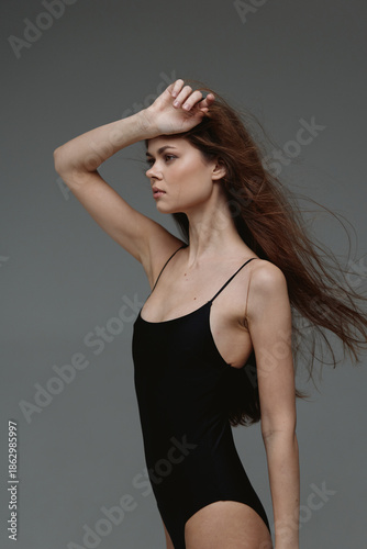 Slim woman in black bodysuit posing with hand on forehead against grey background, contemplative mood, studio fashion shot
