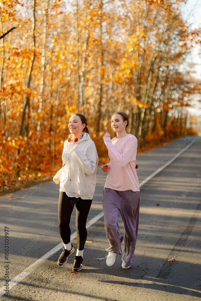 Fototapeta premium Two friends jog along a tree-lined road, surrounded by vibrant fall foliage. Their laughter fills the air as they share a fun moment on a bright, sunny day.