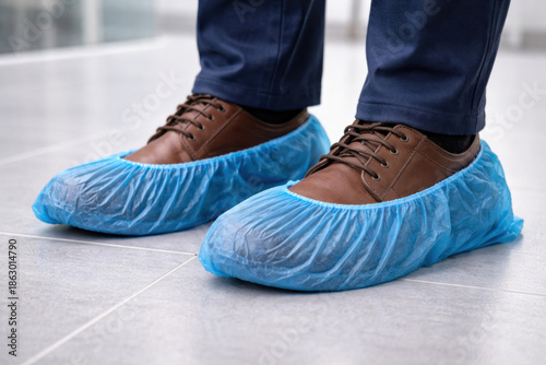 Feet in brown shoes with blue disposable shoe covers on a clean floor, illustrating hygiene and safety in public or medical spaces.