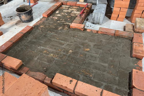 Fresh brickwork of a heating stove with a bench base. Laid out of brick on a concrete screed. Bricks and tools nearby.