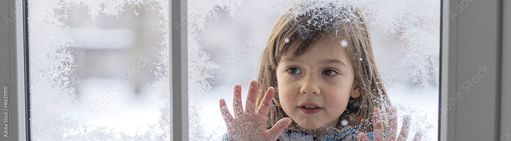Naklejka premium young child face peeks behind frosty windowpane mesmerized falling snowflakes soft warm natural light subtle