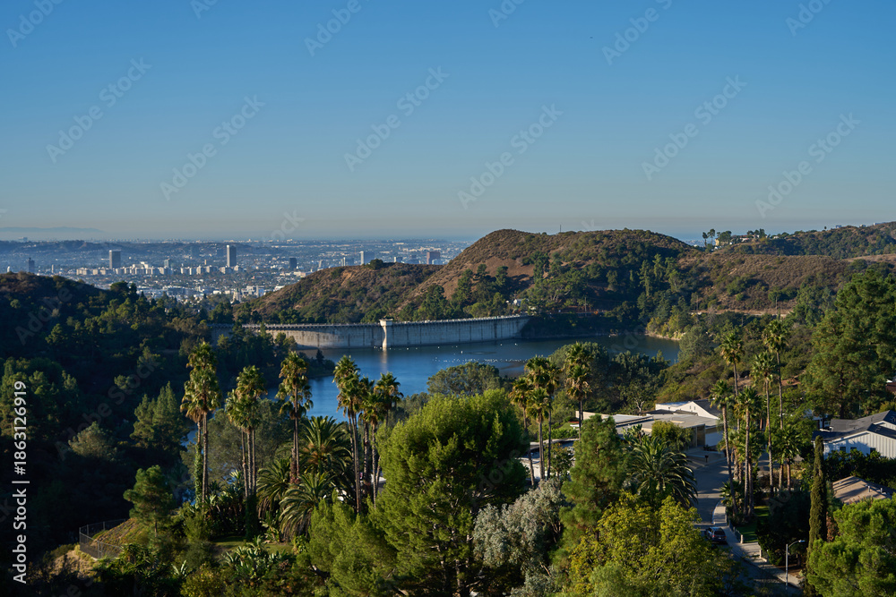 Obraz premium Los Angeles, USA - October 18, 2025 - Hollywood Reservoir dam and city skyline view with lush green trees and palm trees
