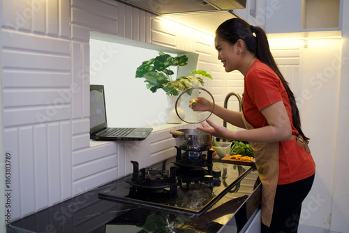 Принти на полотні A young Asian mother is learning to cook by watching a cooking tutorial on her laptop while opening the lid of the pan and inhaling the aroma of the cooking