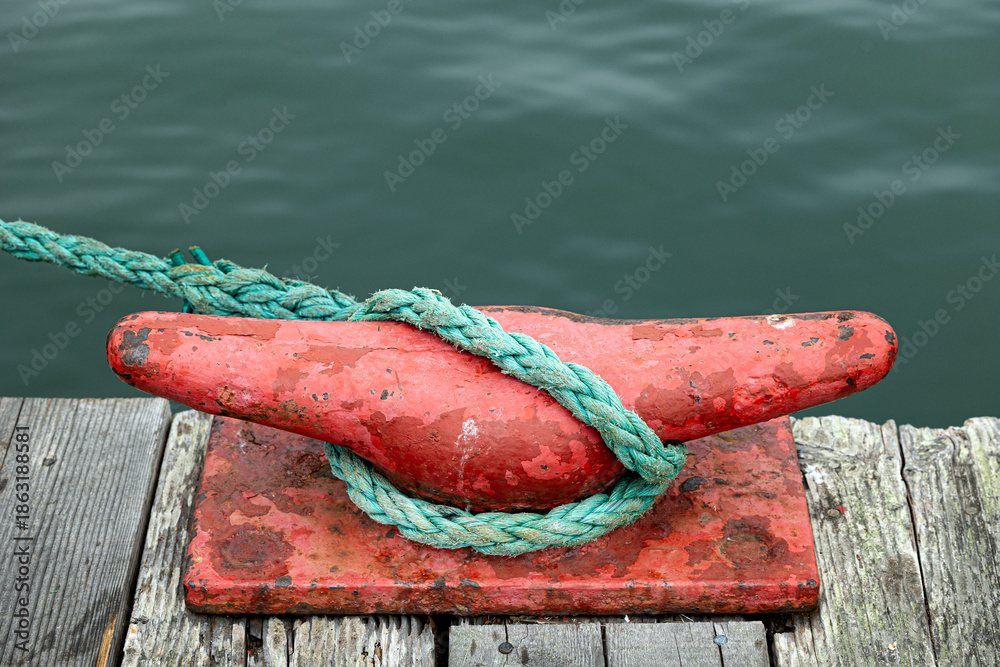 Fototapeta premium Close-up of a weathered red metal dock cleat wrapped with green rope on a wooden pier beside calm water. Nautical detail showing texture, wear, and marine elements, suitable for boating, maritime, har
