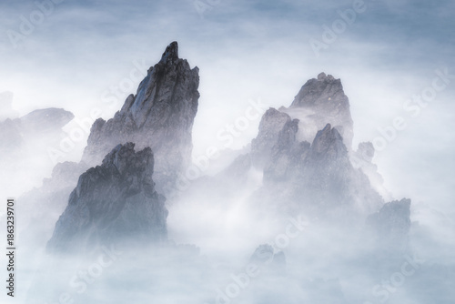 Long exposure landscape shot featuring jagged dark rock formations emerging from dense white mist or sea spray. High-contrast, minimalist aesthetic for editorial use.