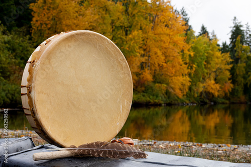 A close up image of a hand made leather meditation drum with a sacred feather and colorful autumn leaves reflecting off clear lake waters in the background. 