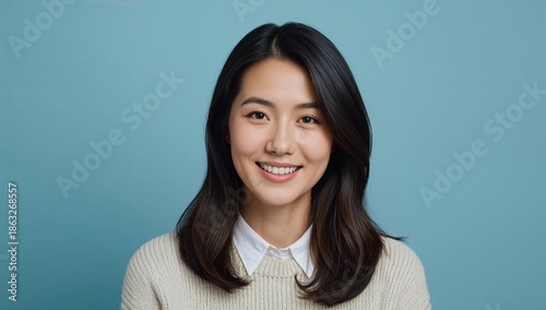 Smiling Woman With Dark Hair and Light Sweater Stands in Front of Blue Backgr...