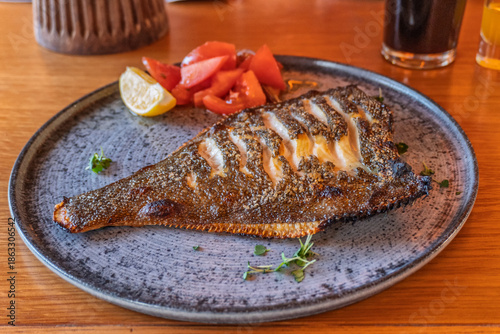 Obraz na płótnie A traditional dish of the Baltic region, grilled flounder is golden-brown and seasoned with spices, served on a ceramic plate with fresh tomatoes, a lemon wedge, and herbs