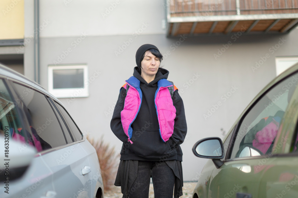 Fototapeta premium Young person in bright vest standing between parked cars, carrying backpack and beanie, confident urban commuter ready for morning route, candid street style, cool color palette