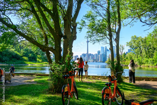 Visitors standing at a shoreline viewpoint on the Toronto Islands, looking across calm water toward the downtown Toronto skyline on a clear summer day.
