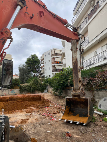 Excavator digging foundation at an urban construction site between residential buildings.Heavy machinery, earthworks and groundwork for infrastructure, housing utility installation in city environment