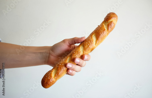 Female hand holds long, golden brown sprinkled with sesame seeds against plain white background. Crust shows distinct score marks, crisp texture. Classic French bread offers fresh baked appearance,