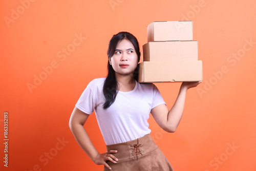 Young woman holding a stack of boxes, looks tired with a hand on her hip and balancing the heavy load on one hand. isolated on orange background